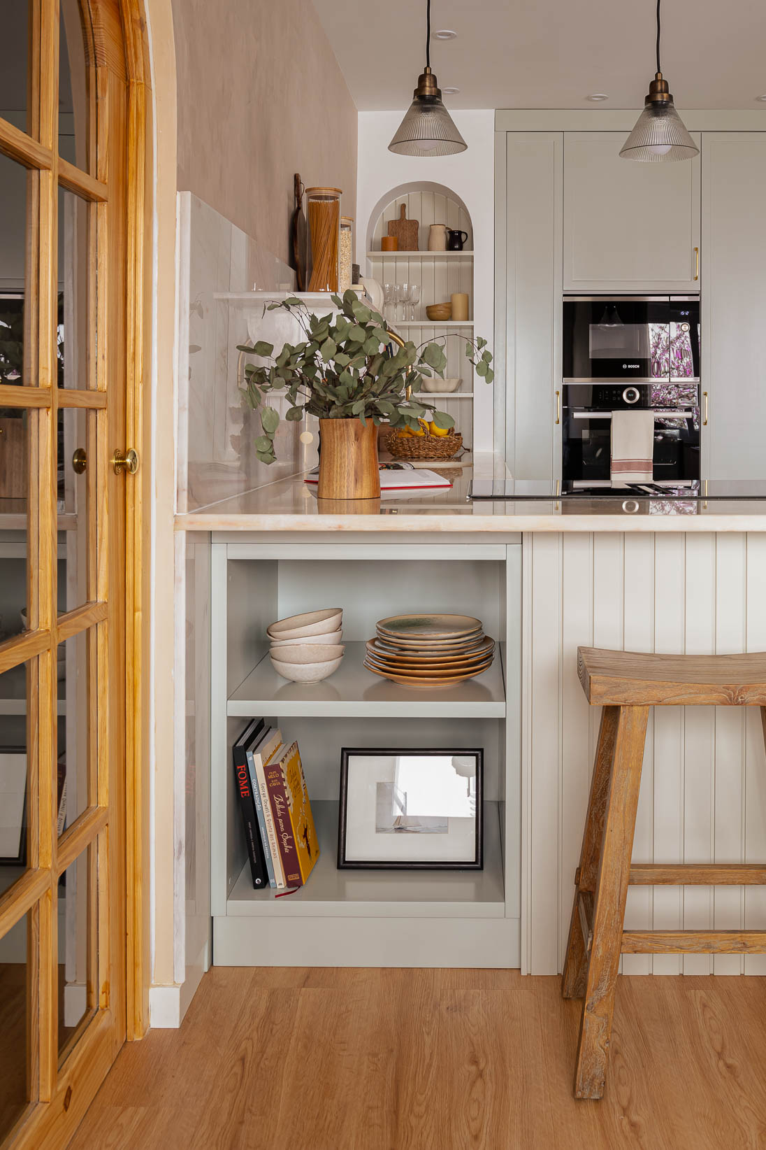 Casa do Paço - Cozinha aberta com banco de madeira, e bancada em mármore claro, móveis ripados a vere/sage green, com arrumação aberta. Backsplash a mármore branco; estante em mármore e hardware em metal dourado. Dispensa com porta de madeira e vidro