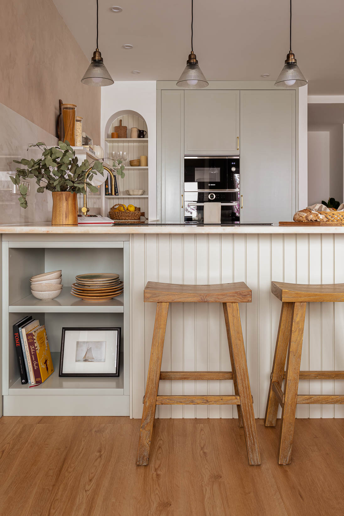 Casa do Paço - Cozinha aberta com banco de madeira, e bancada em mármore claro, móveis ripados a vere/sage green, com arrumação aberta. Backsplash a mármore branco; estante em mármore e hardware em metal dourado. Dispensa com porta de madeira e vidro. Arco decorativo para funcioanr como estante