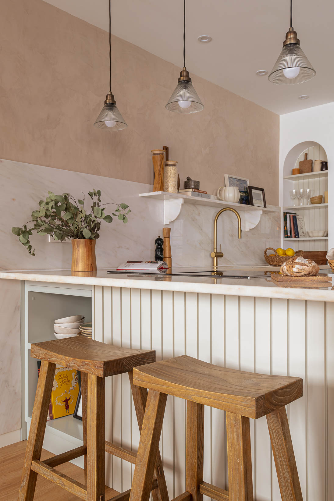 Casa do Paço - Cozinha aberta com banco de madeira, e bancada em mármore claro, móveis ripados a vere/sage green, com arrumação aberta. Backsplash a mármore branco; estante em mármore e hardware em metal dourado. Dispensa com porta de madeira e vidro; arco decorativo para funcioanr como estante