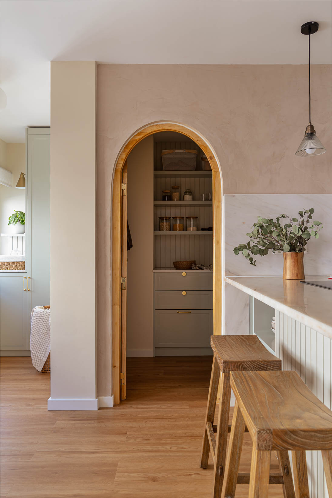 Casa do Paço - Cozinha aberta com banco de madeira, e bancada em mármore claro, móveis ripados a vere/sage green, com arrumação aberta. Backsplash a mármore branco; estante em mármore e hardware em metal dourado. Dispensa com porta de madeira e vidro