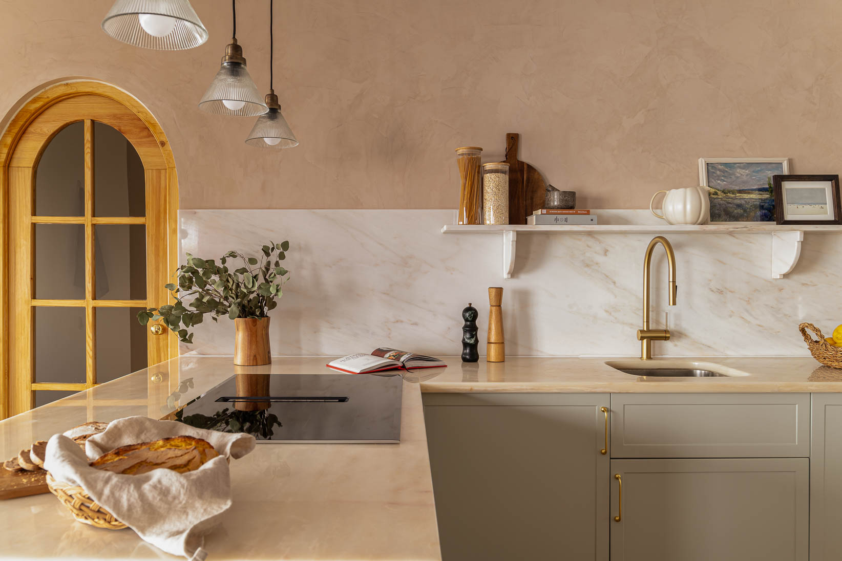 Casa do Paço - Cozinha aberta com banco de madeira, e bancada em mármore claro, móveis ripados a vere/sage green, com arrumação aberta. Backsplash a mármore branco; estante em mármore e hardware em metal dourado. Dispensa com porta de madeira e vidro