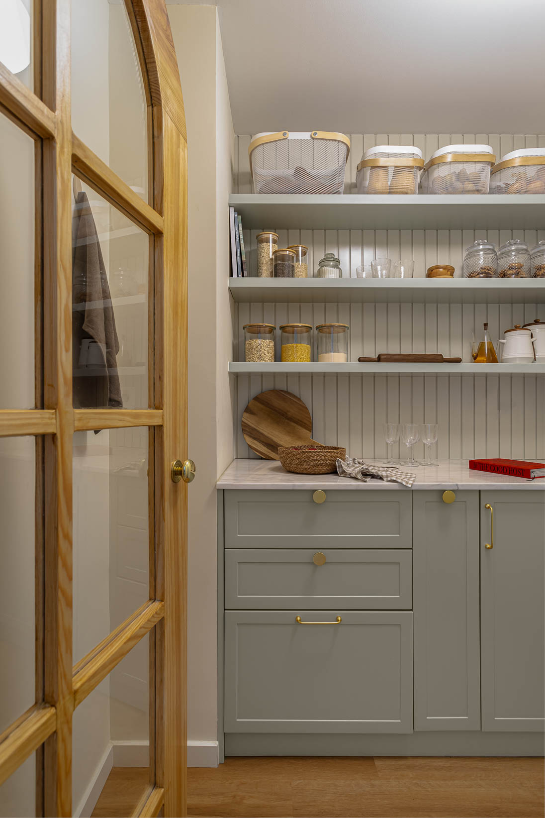 Casa do Paço - Cozinha aberta com banco de madeira, e bancada em mármore claro, móveis ripados a vere/sage green, com arrumação aberta. Backsplash a mármore branco; estante em mármore e hardware em metal dourado. Dispensa com porta de madeira e vidro; frascos de vidro com bolachas e acessórios de cozinha