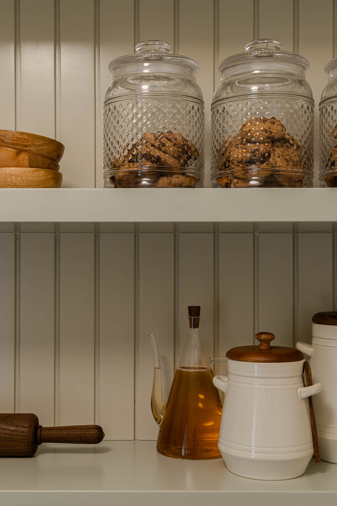 Casa do Paço - Cozinha aberta com banco de madeira, e bancada em mármore claro, móveis ripados a vere/sage green, com arrumação aberta. Backsplash a mármore branco; estante em mármore e hardware em metal dourado. Dispensa com porta de madeira e vidro; frascos de vidro com bolachas e acessórios de cozinha