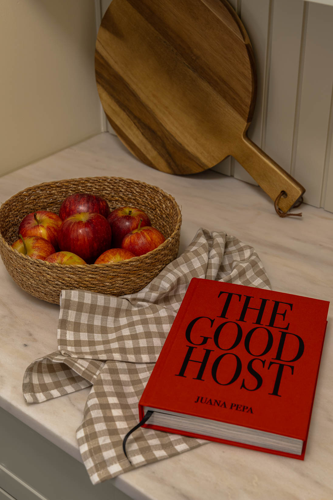 Casa do Paço - Cozinha aberta com banco de madeira, e bancada em mármore claro, móveis ripados a vere/sage green, com arrumação aberta. Backsplash a mármore branco; estante em mármore e hardware em metal dourado. Dispensa com porta de madeira e vidro; frascos de vidro com bolachas e acessórios de cozinha; pano de cozinha, cesto da fruta e tábua de madeira